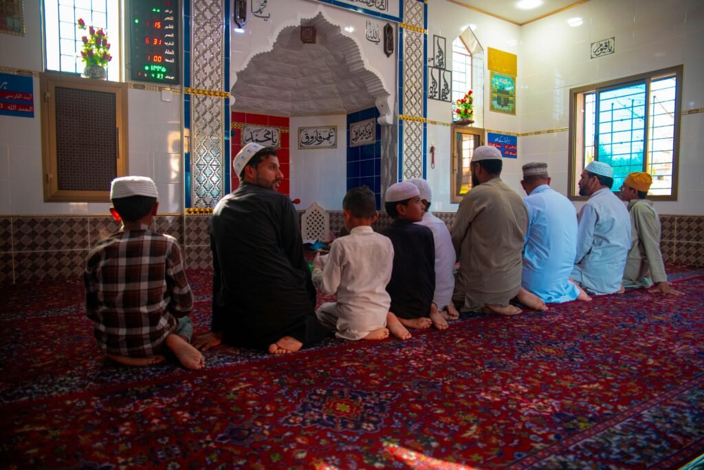 Men and boys kneeling on a colorful carpet, praying in a mosque with sunlight filtering through windows.